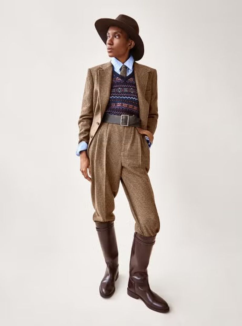 A person in a tweed suit, sweater vest, hat, and riding boots poses in a fashion editorial shot against a neutral background.
