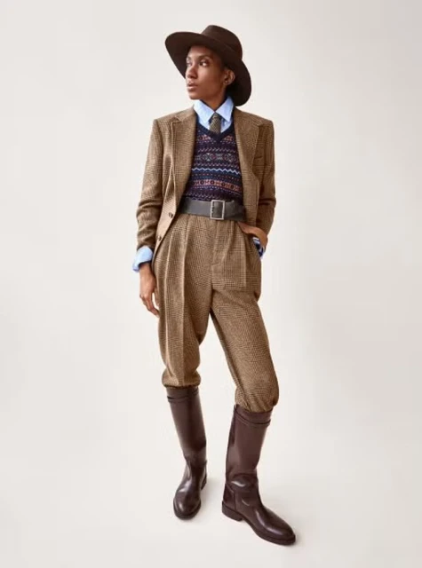 A person in a tweed suit, sweater vest, hat, and riding boots poses in a fashion editorial shot against a neutral background.