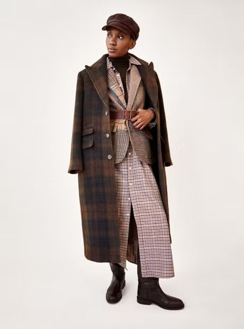 Woman poses in a brown plaid overcoat, blazer, and a matching dress with black leather boots on a white studio backdrop.