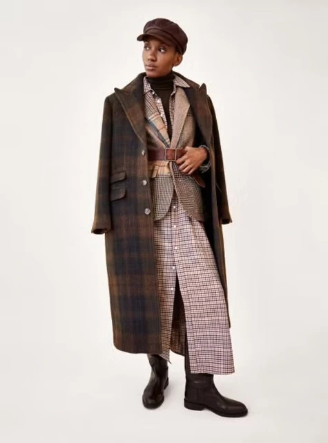 Woman poses in a brown plaid overcoat, blazer, and a matching dress with black leather boots on a white studio backdrop.