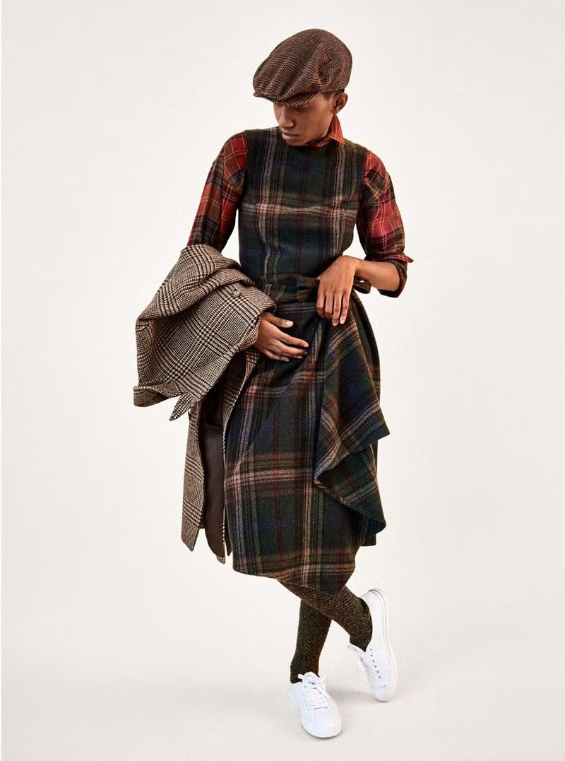 Full-length shot of a woman in a plaid dress, newsboy cap, and sneakers, holding a tweed coat against a plain studio background.
