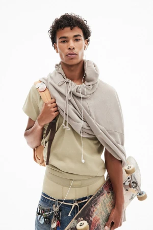 Young adult male fashion model poses in a pale green outfit with a skateboard in front of a white background in a studio shot.