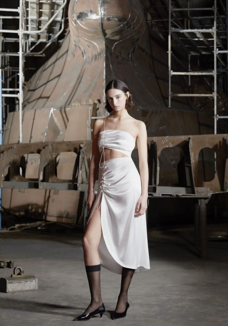 Fashion model in white two-piece outfit poses indoors against an industrial backdrop, wearing sheer black socks and black heeled shoes.