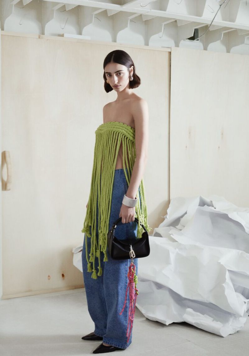 Brunette woman poses in strapless fringed top and wide-leg jeans, holding black purse against a background of white crumpled paper.
