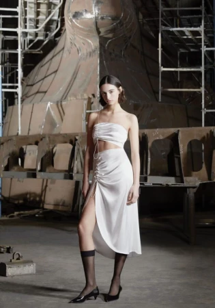 Model posing in a white ruched satin two-piece skirt set and black pointed heels inside an industrial shipyard workshop.