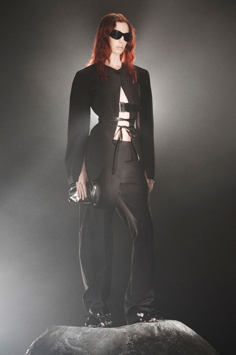 Woman in black jacket, pants, and sunglasses stands on a rock-like surface against a dark background, holding a silver clutch.