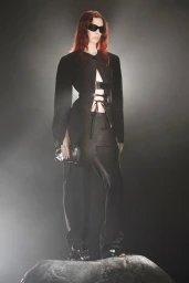 Model with reddish hair in black cutout blazer, trousers, and dark sunglasses posing atop a rock in misty lighting.
