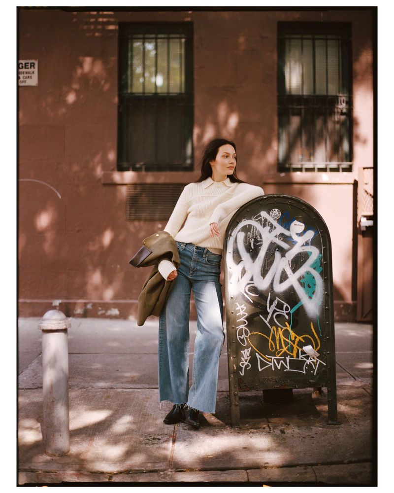 Adult woman stands in urban setting near graffiti-covered object. The woman wears jeans and a sweater, and carries a coat and cup.