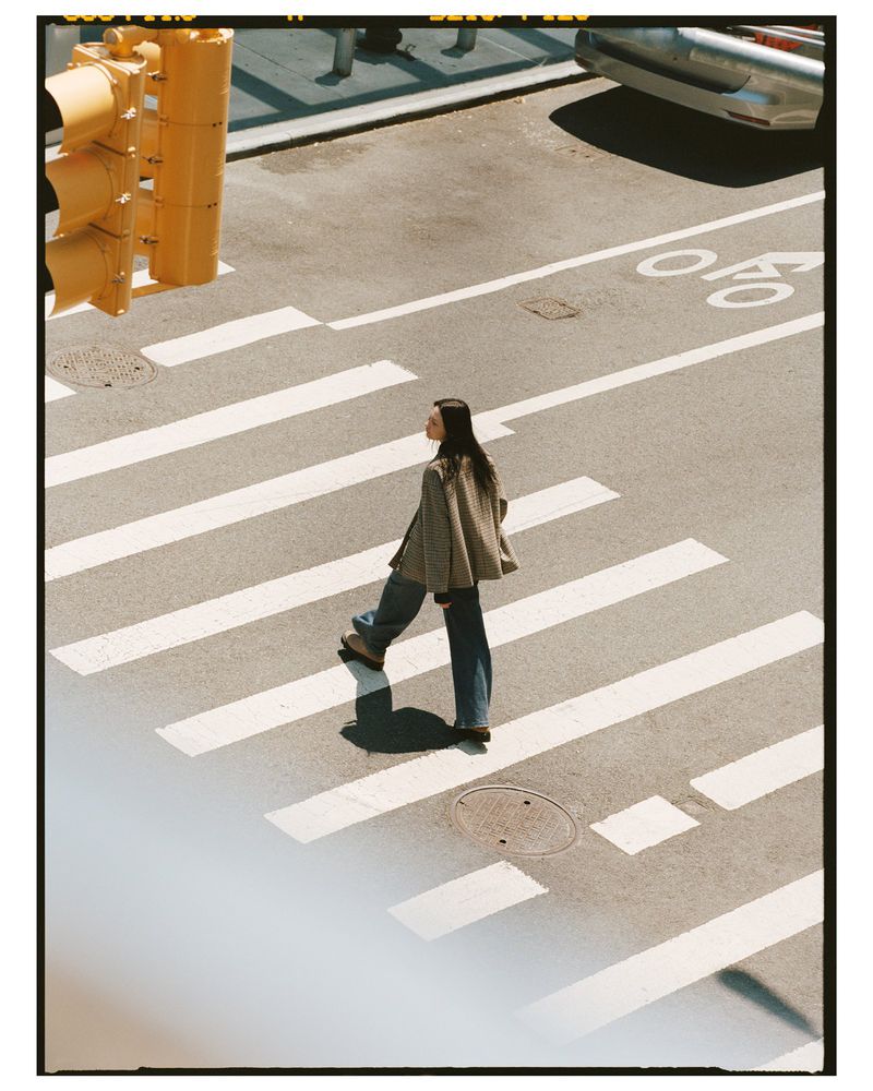 Woman in a plaid jacket walks across a white-striped crosswalk in an urban setting on a sunny day with a traffic light overhead.