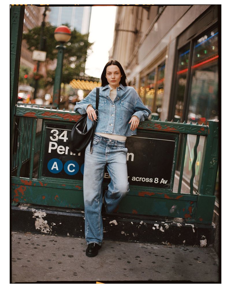 Woman in denim jacket and jeans posing in front of a subway entrance in an urban setting with a city street visible in the background.