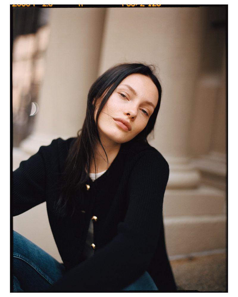 Portrait of a brunette woman in a black cardigan and jeans, posing outdoors against an off-white stone wall with natural light.