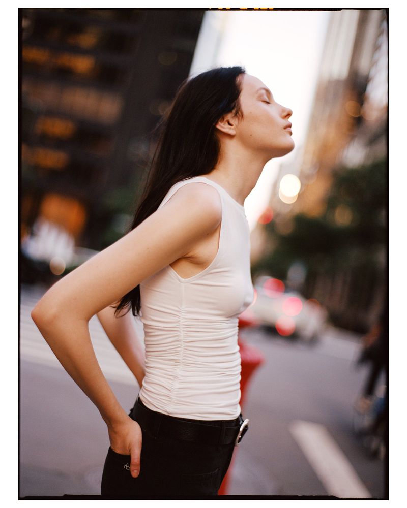 Young woman in a white top and black pants, stands outside with eyes closed, enjoying the sunlight against a city backdrop.