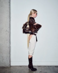 Model Emily Fay posing in profile wearing a brown leather jacket, cutout white pants, and heeled boots against a white wall.