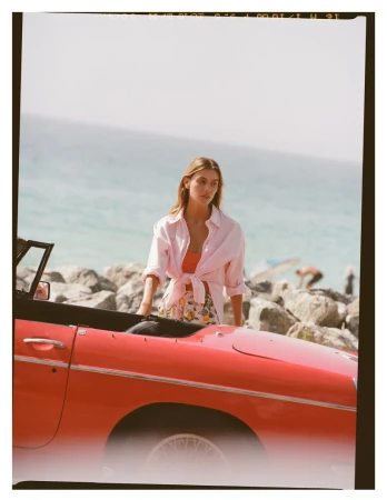Woman in a pink shirt and floral shorts standing next to a red convertible car by the beach, with ocean in the background.