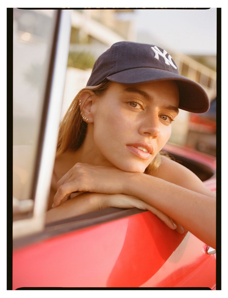 Close-up portrait of a young adult female wearing a baseball cap, posing with her arms resting on the door of a red car.