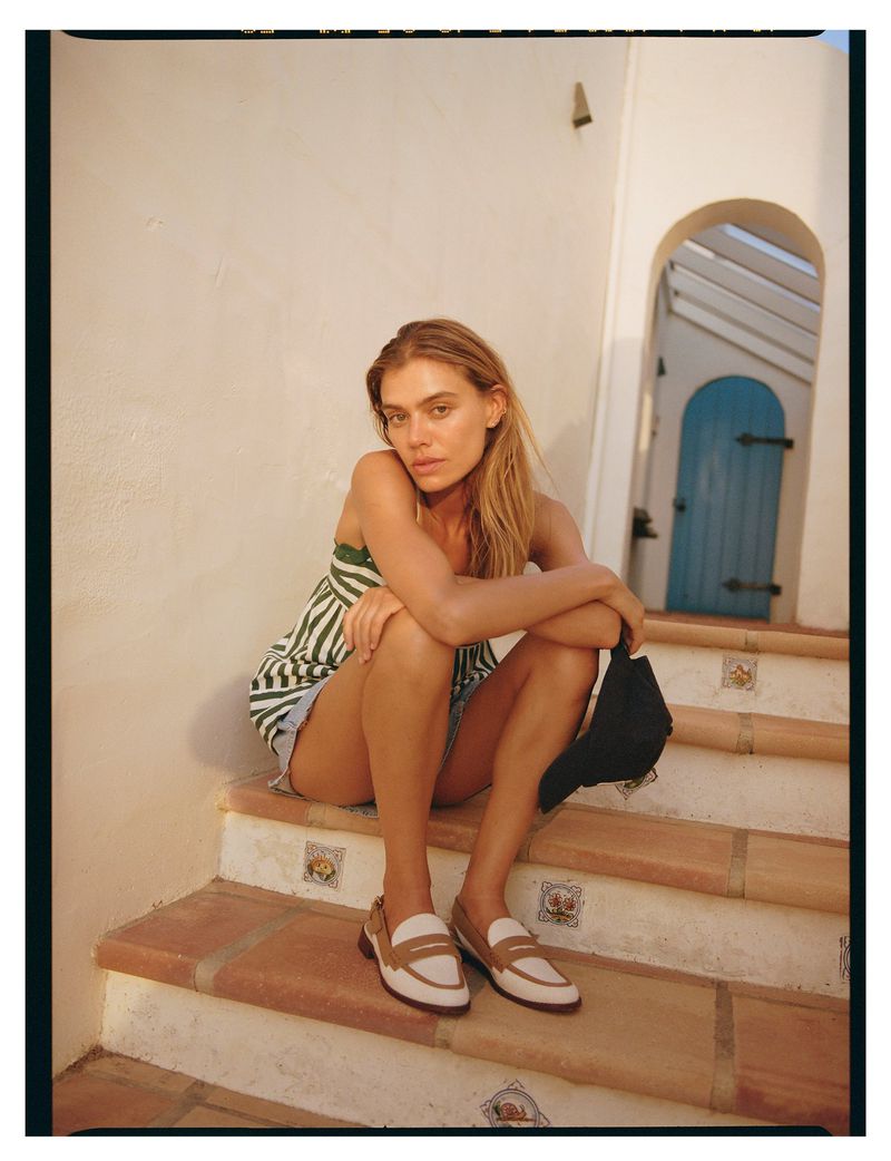 Young adult woman with blonde hair sits on outdoor tile steps in casual summer clothes and leather loafers holding a black cap.