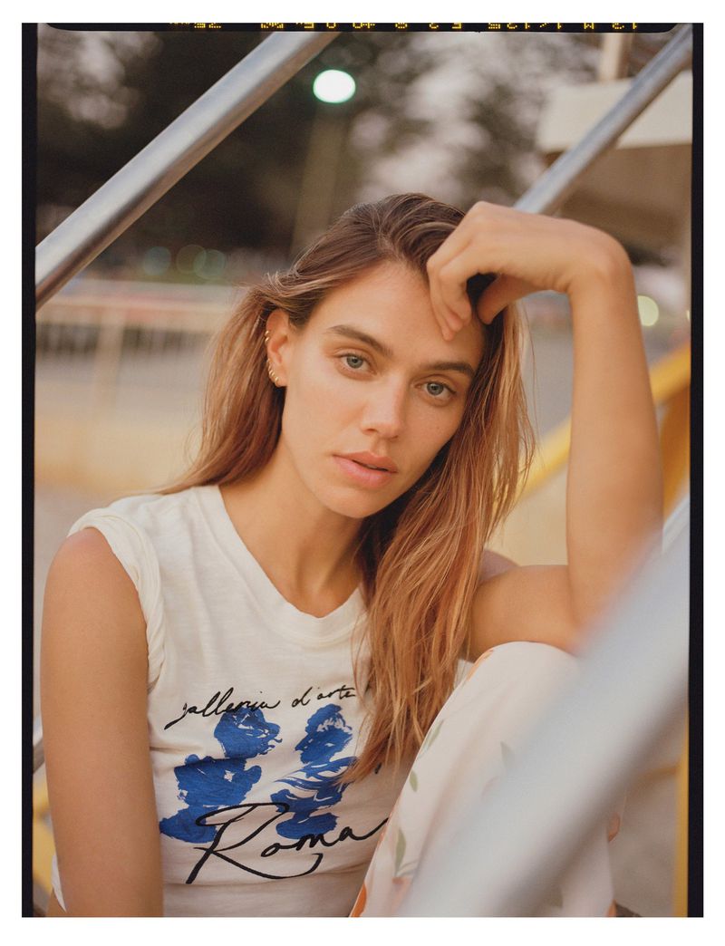 Portrait of a young woman with long light-brown hair, wearing a graphic t-shirt, resting her hand on her forehead on a stairwell.