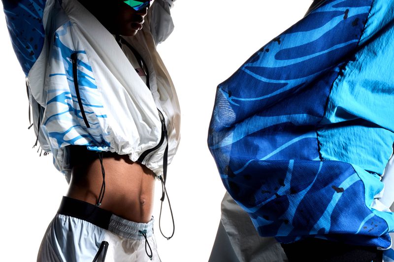 Stylish person in light colored windbreaker and shorts against white backdrop. Abstract blue wave designs accent jacket. 