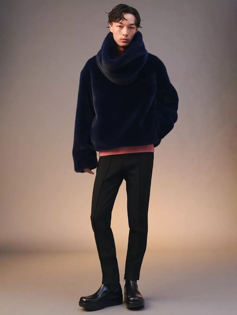 Full-length shot of a young adult male in a navy faux fur coat, black pants, and boots, posing in front of a gradient beige background. 