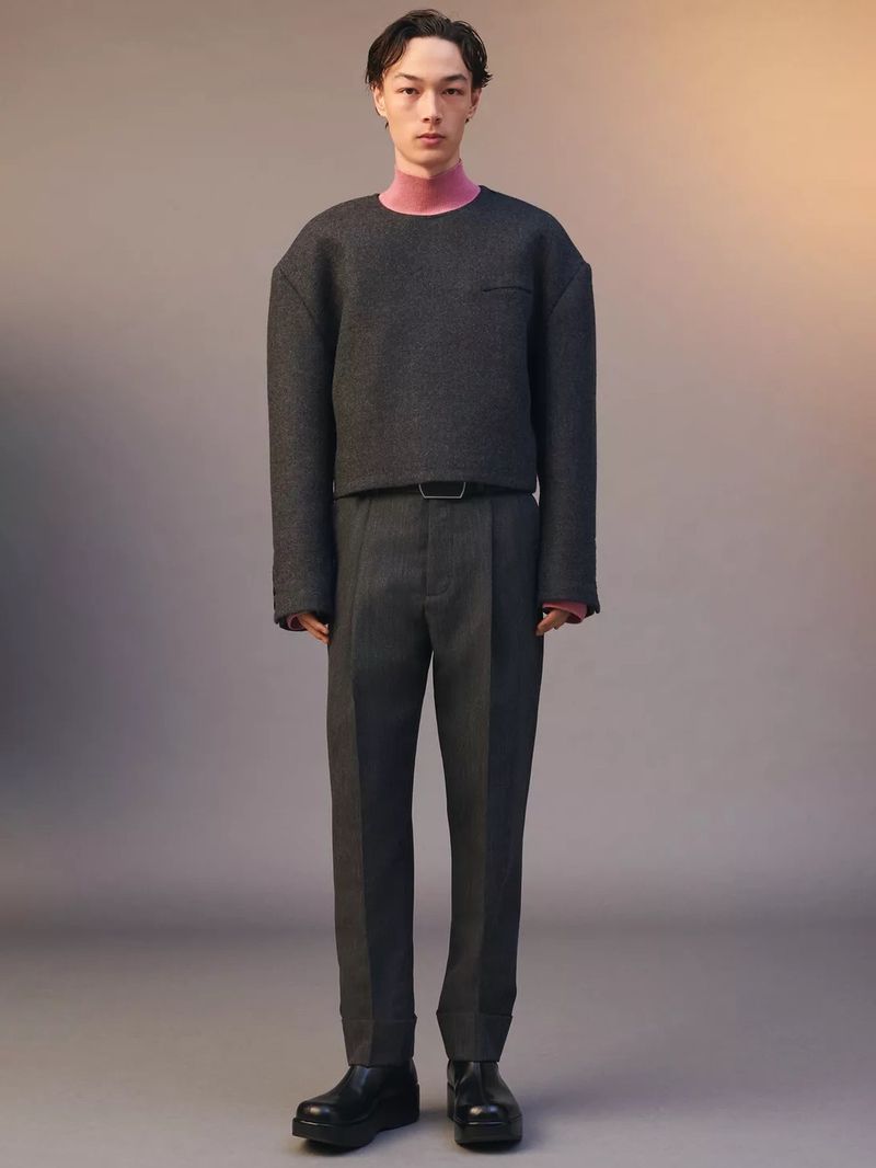Full-length studio shot of an adult male fashion model wearing a grey sweater, pink turtleneck, grey pants, a black belt, and black shoes.
