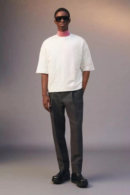 Full-length shot of a slender, dark-skinned man wearing a white short-sleeve t-shirt, grey pants, and black chunky shoes in a studio setting.