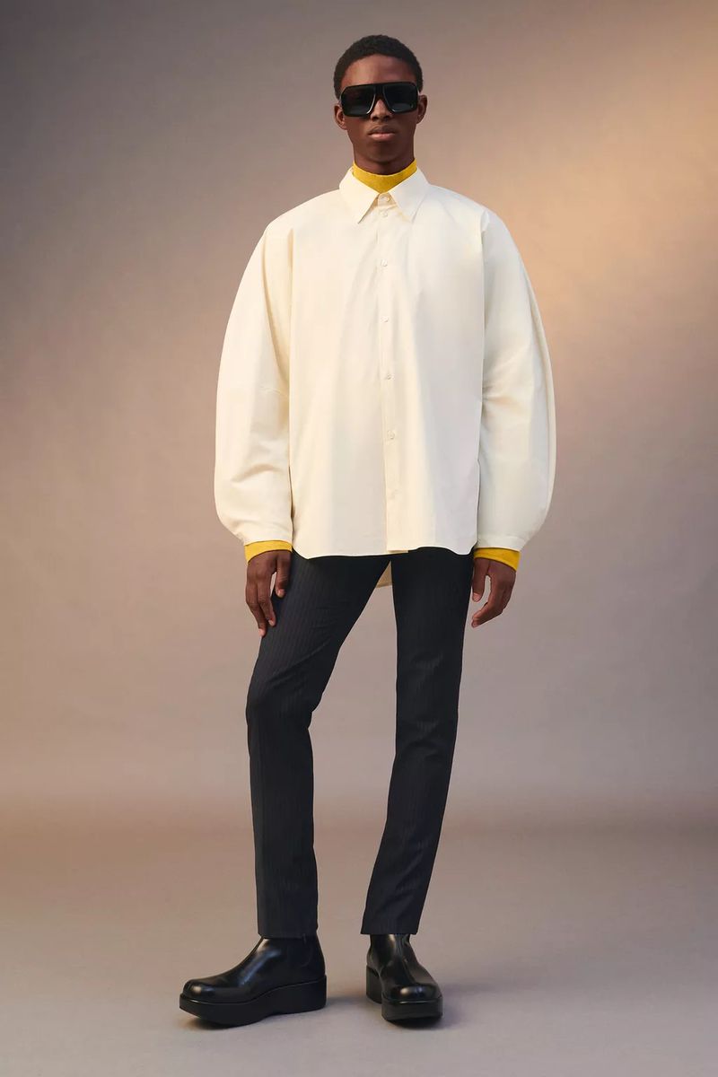 Fashion model poses in an off-white button-down shirt, black trousers, and boots against a soft gradient backdrop in a studio setting.