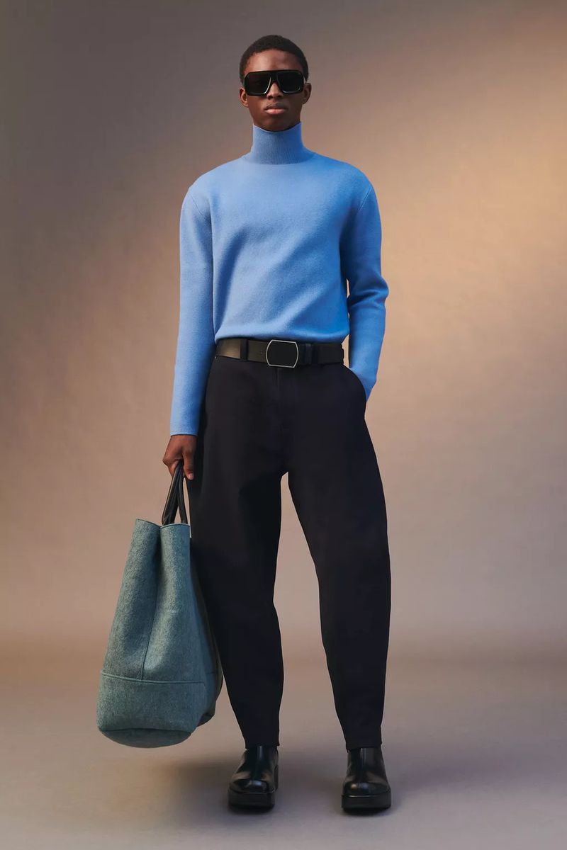 Model wearing a blue turtleneck, black trousers, and sunglasses holding a teal tote bag against a gradient background.