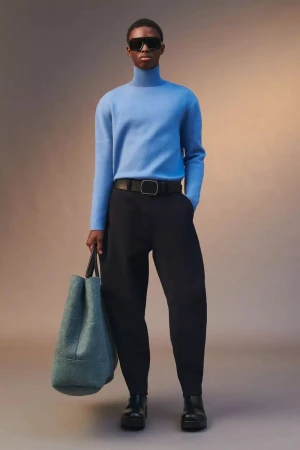 Model wearing a blue turtleneck, black trousers, and sunglasses holding a teal tote bag against a gradient background.