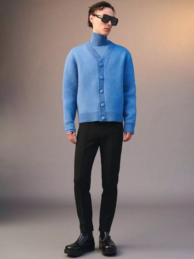 Full-length portrait of a man in a blue cardigan, black pants, and black boots. He is standing in a neutral studio background.