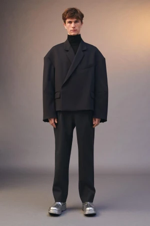 A single male model stands in a photo studio wearing an oversized black blazer, black turtleneck, black pants, and silver shoes.