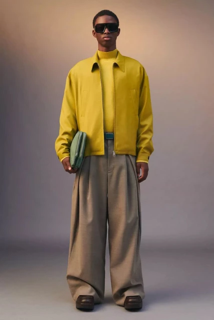 Man stands in neutral studio wearing yellow shirt and jacket with large khaki pants, holding green clutch, with sunglasses and brown shoes.