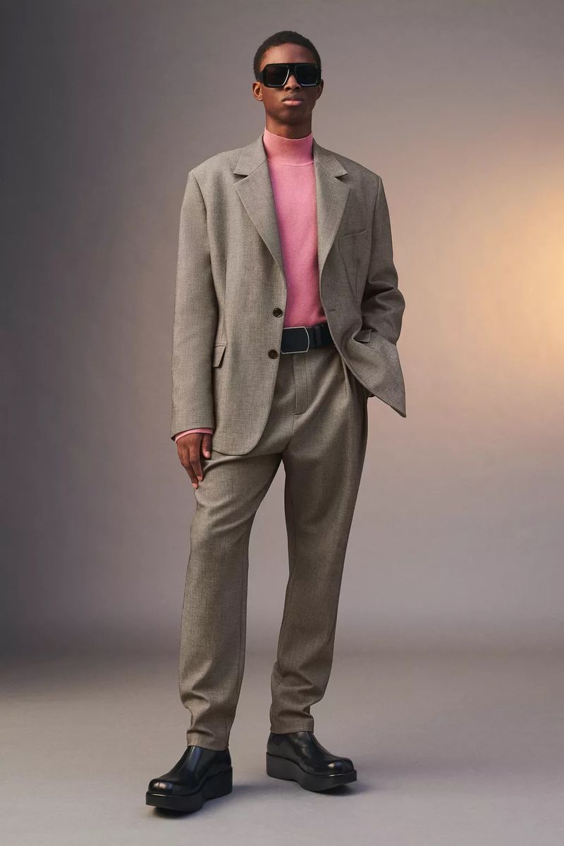Man in a tan suit and sunglasses standing against a gray background with a subtle light gradient, creating a sophisticated fashion image.