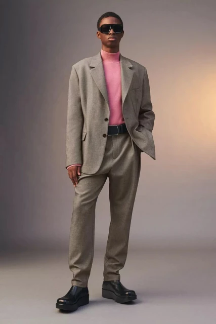 Man in a tan suit and sunglasses standing against a gray background with a subtle light gradient, creating a sophisticated fashion image.