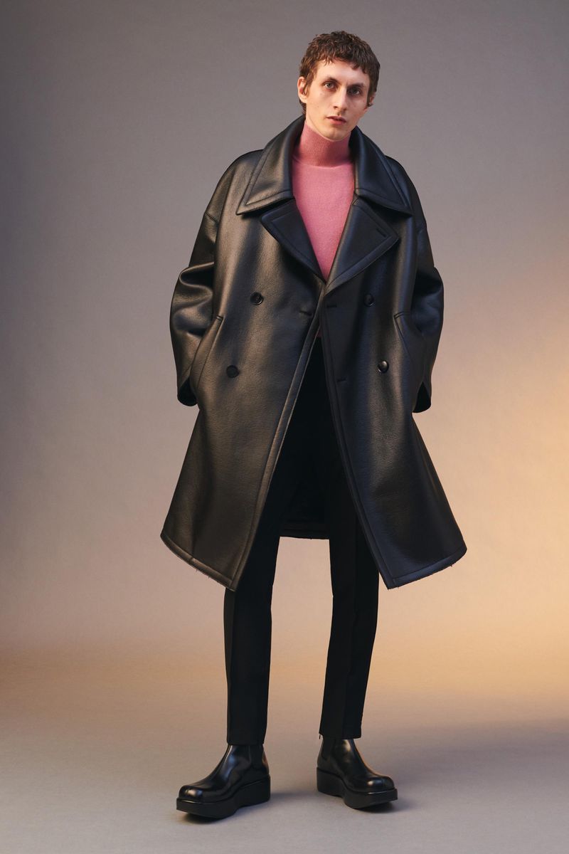 Full-length studio portrait of adult male fashion model in a black leather trench coat, pink turtleneck, and black pants and dress shoes.