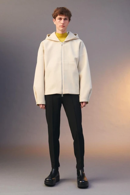 Full-length portrait of a young adult man wearing a hooded jacket, turtle neck, fitted pants, and platform boots in a studio setting.