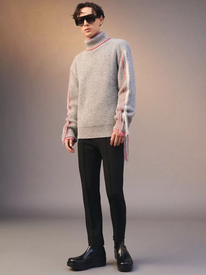 Man in gray turtleneck sweater with fringe details, black pants, and black boots poses in a studio with a neutral-toned background.