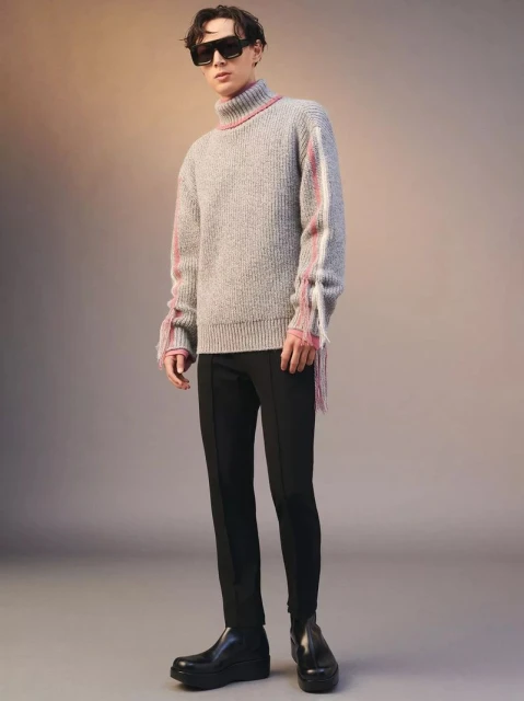 Man in gray turtleneck sweater with fringe details, black pants, and black boots poses in a studio with a neutral-toned background.
