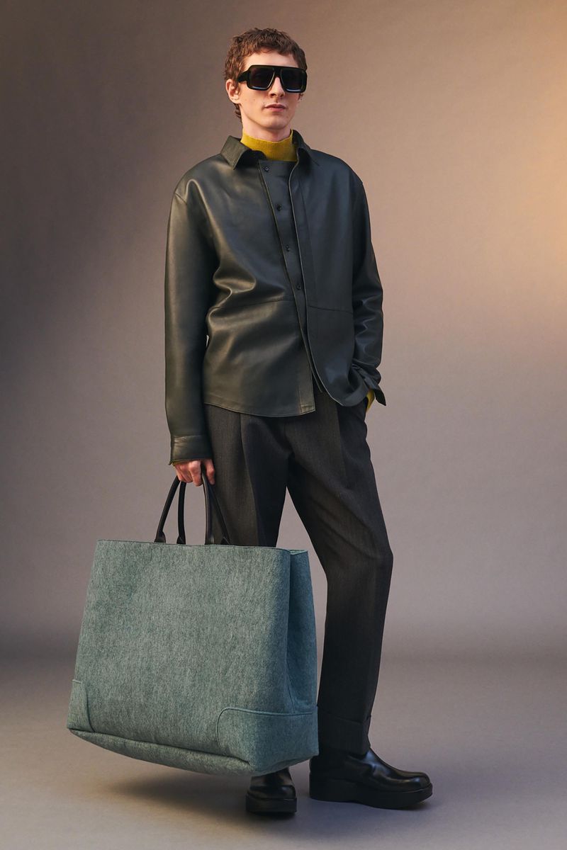 Studio shot of an adult male fashion model wearing sunglasses, black leather shirt, and holding a large tote bag, against a grey background.