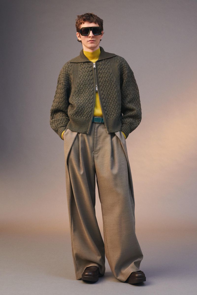 Male model wearing a green knit zip-up sweater, yellow turtleneck, and beige pleated trousers stands in a studio setting.