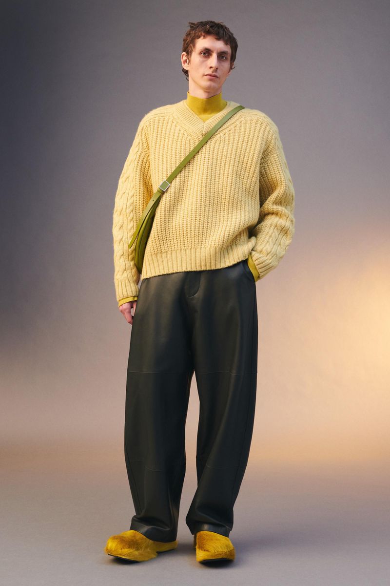 A young adult male poses in a studio wearing a yellow sweater, yellow turtleneck, green shoulder bag, black leather pants, and yellow slippers.