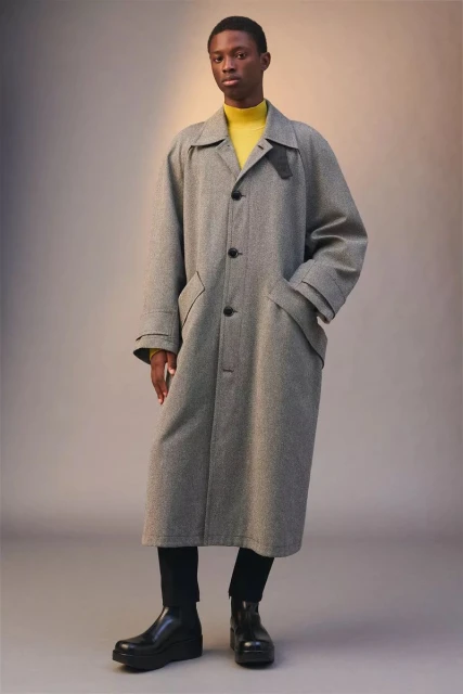 A fashionable Black male model poses in a grey trench coat, yellow turtleneck, black pants, and platform boots against a neutral backdrop.