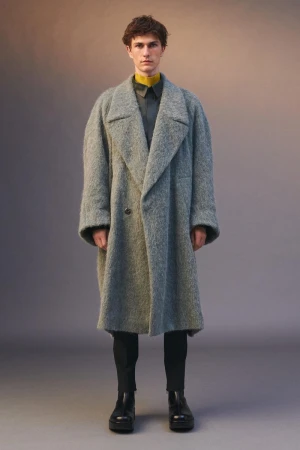 Model standing facing forward wearing an oversized grey textured coat over dark trousers and black leather shoes against a studio backdrop
