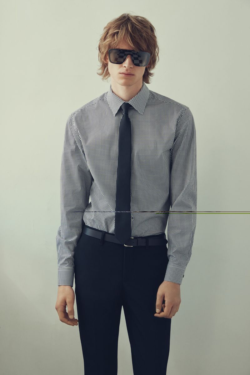 Male model in checkered shirt, black tie, and sunglasses poses against a neutral backdrop for a fashion editorial shot in a photo studio.