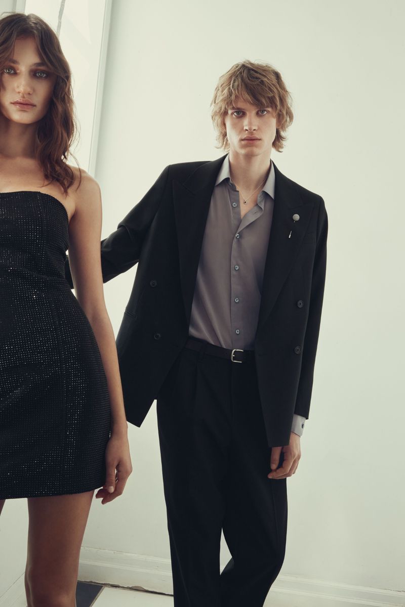 A dark-haired woman in a black dress and a blond man in a grey shirt and black suit pose in front of a white wall.