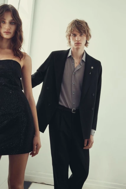 A dark-haired woman in a black dress and a blond man in a grey shirt and black suit pose in front of a white wall.