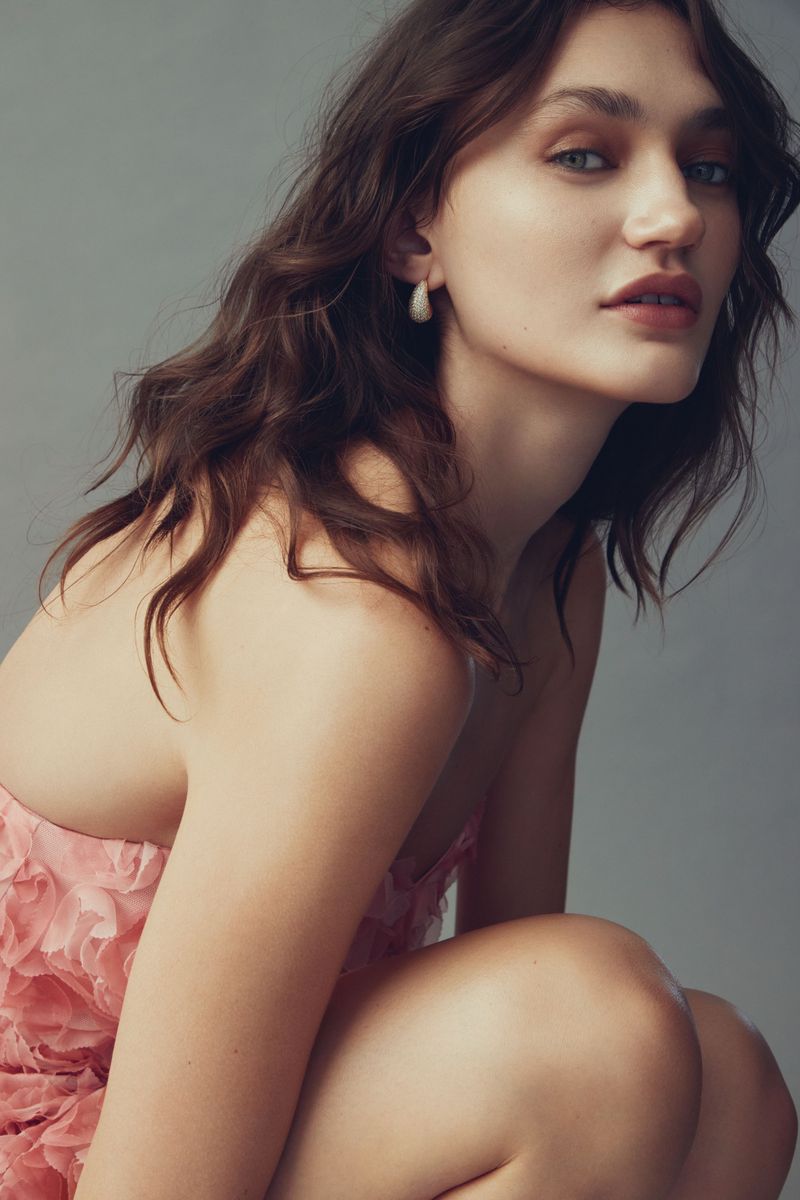 Portrait of an adult woman with brown wavy hair, wearing a pink dress, posing in front of a grey background.