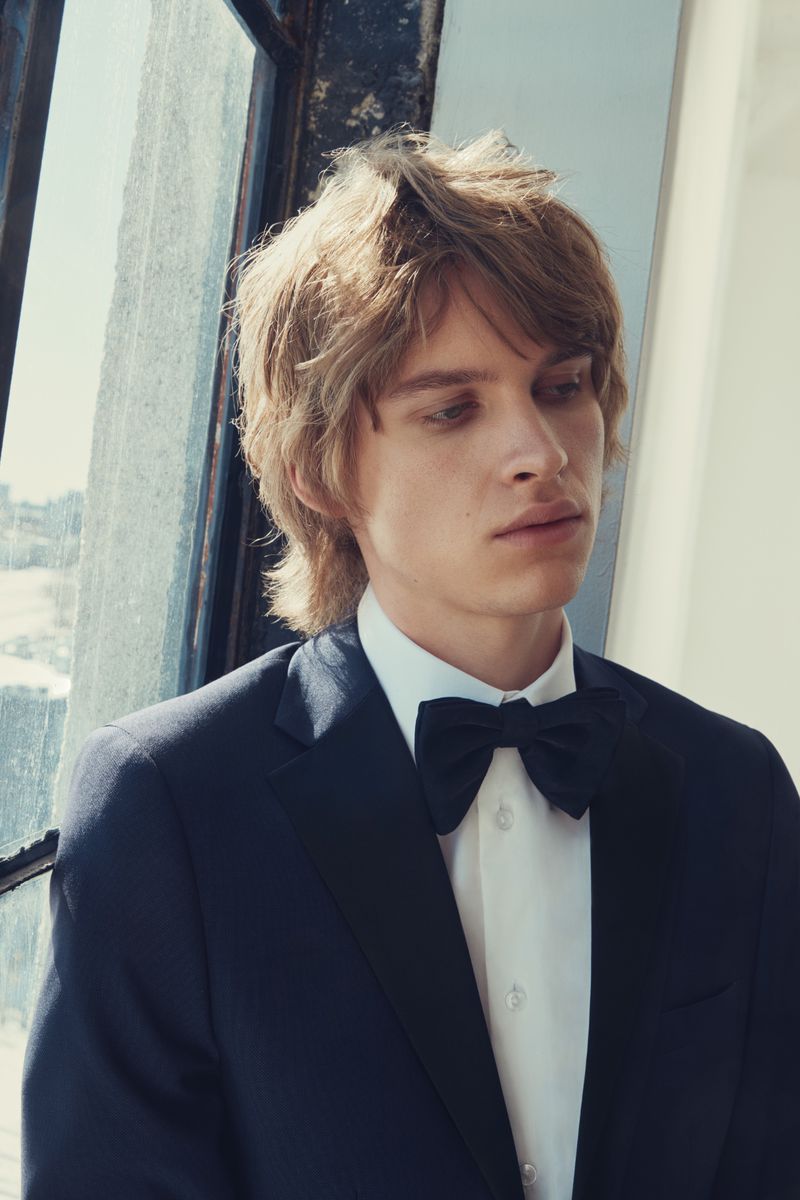 Young blond male in formal attire, including a suit jacket, white shirt, and bow tie, posing near a sunlit window.