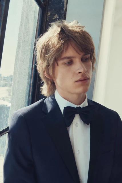 Young blond male in formal attire, including a suit jacket, white shirt, and bow tie, posing near a sunlit window.
