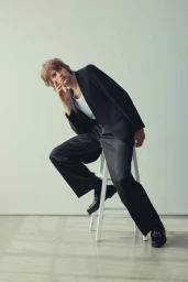 Model Parker Olson sits on a white stool wearing a black tailored suit, wide-leg trousers, and white tank top in a bright studio.