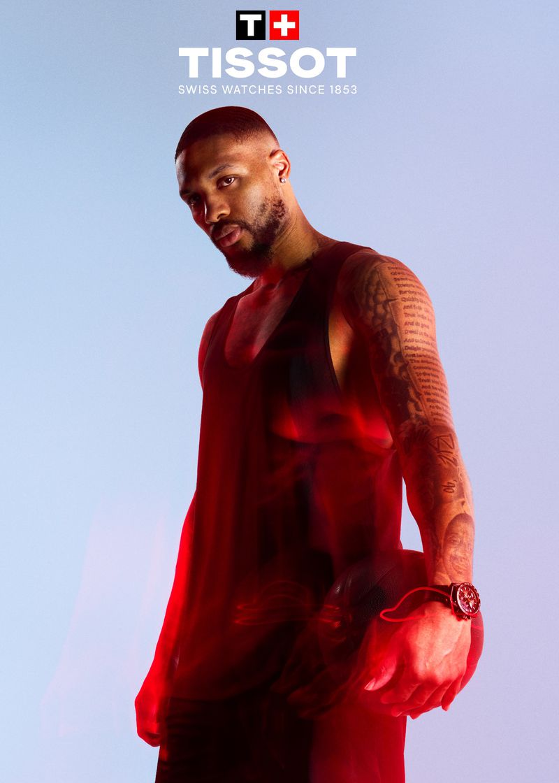 Portrait of adult male athlete with tattoos wearing a watch, posing against a light blue background with a red light effect.